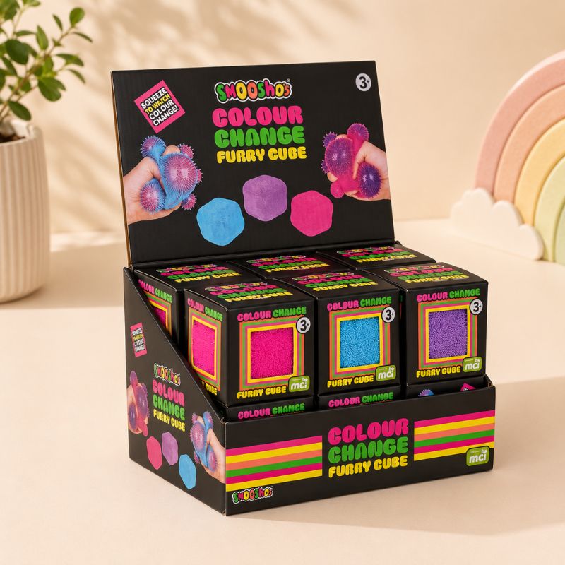 A retail display box filled with SmooSho Colour Change Furry Cube toys, shown on a warm beige tabletop with a potted plant and pastel rainbow decor in the background.