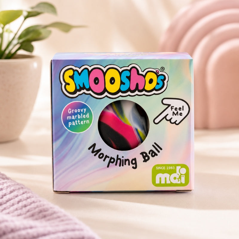 A boxed squishy toy called SMOOOSHs Morphing Ball, shown in its retail packaging on a soft pastel surface with a plant and pink decor blurred in the background. The box is iridescent with colorful branding and a circular window revealing the marbled ball inside.