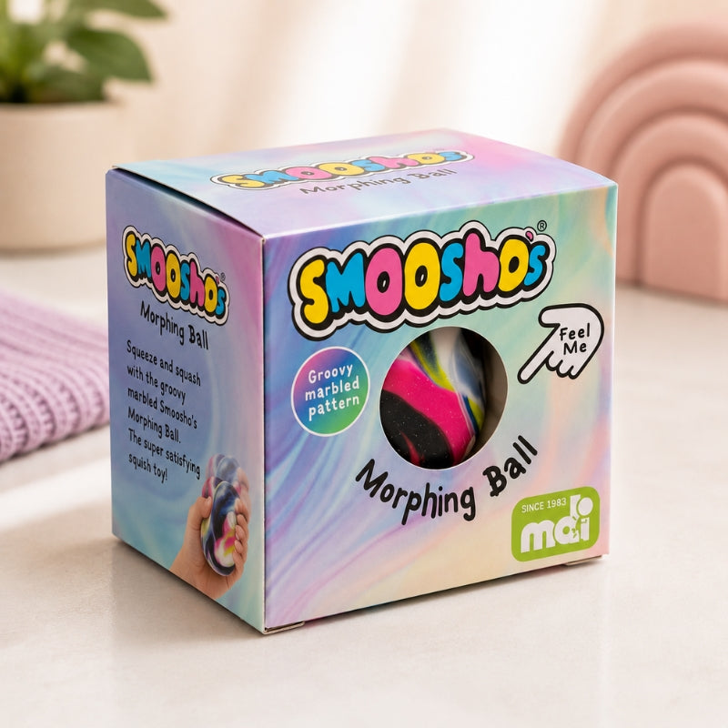 A boxed Smooshos Morphing Ball toy in pastel iridescent packaging, photographed on a light tabletop with a soft blurred background. The box shows the Smooshos logo, a circular window revealing the pink-and-black morphing ball, and text including 'Groovy marbles pattern' and 'Feel Me'.