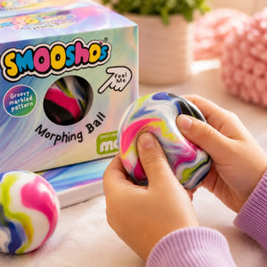 A person's hands squeezing a colorful marbled 'Morphing Ball' stress toy, with its branded retail box in the background and another ball resting nearby on a soft tabletop.