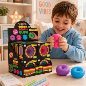 A smiling boy in a light blue sweatshirt holds a pink glow-in-the-dark donut-shaped toy while standing next to a black retail display box of 'Gooberz Super Squeeze Glow' donuts. Two more glow donuts, one purple and one blue, rest on the light wood tabletop in front of him, with colorful pencils nearby.