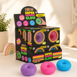 A retail display box of 'Bloomio' Super Squeeze Glow Doughnut candies, with three glow-in-the-dark doughnuts in purple, pink, and blue placed in front on a light-colored surface.