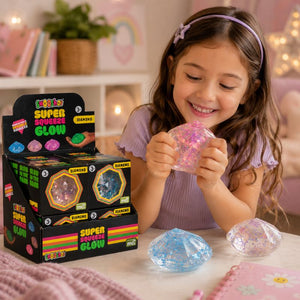 A young girl with a purple headband smiles while holding a pink glittery squeeze gem toy. In front of her are two larger diamond-shaped squeeze gems, one blue and one clear, and a display box of colorful 'Glow Pop' Super Squeeze Glow toys on a table in a cozy, softly lit room.