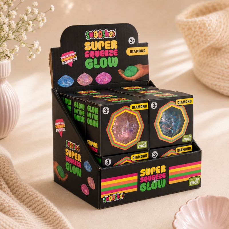 A retail display box filled with Mooshoo Super Squeeze Glow candy boxes, styled on a beige fabric surface with soft decorative props.