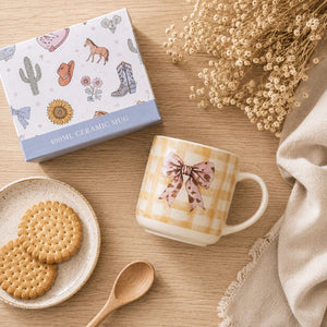 A ceramic Splosh -Saddle & Bloom Bow Mug is shown next to its product box on a wooden table with cookies, a spoon, and dried flowers.