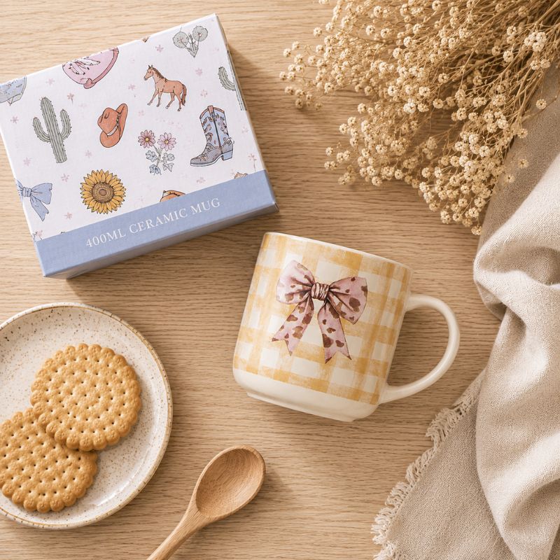 A ceramic Splosh -Saddle & Bloom Bow Mug is shown next to its product box on a wooden table with cookies, a spoon, and dried flowers.