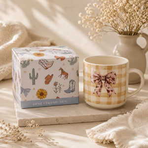 A 400ml Splosh -Saddle & Bloom Bow Mug displayed next to its illustrated gift box on a stone tray with dried flowers and soft textiles in the background.
