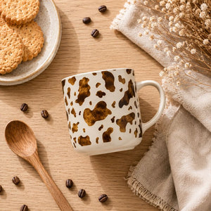A Splosh Saddle & Bloom Cow Mug, styled on a wooden table with coffee beans, a wooden spoon, cookies on a plate, and a beige cloth with dried flowers.