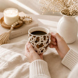 A person holds a Saddle & Bloom Cow Mug filled with black coffee. The cozy scene is styled with a lit candle, a book, and dried flowers in a vase on a warm, sunlit background.
