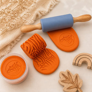 Orange playdough with textured roller, rolling pin, stamp, and wooden toys on a neutral aesthetic background.