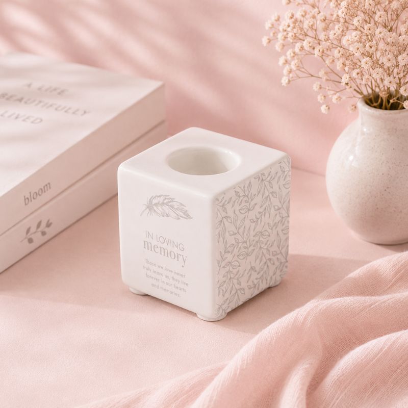 White ceramic candle holder with 'In Loving Memory' text on a pink surface with a vase of flowers in the background.