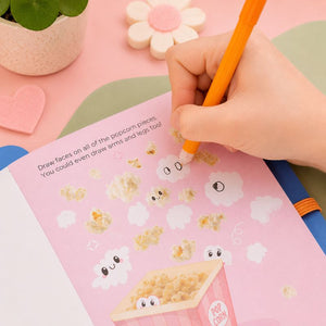 Hand holding a pencil over a pink activity book with cartoon popcorn and clouds, on a colorful surface.
