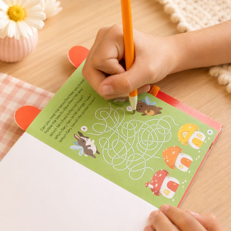 Child's hand holding an orange pencil over a coloring book with a maze on a wooden table.