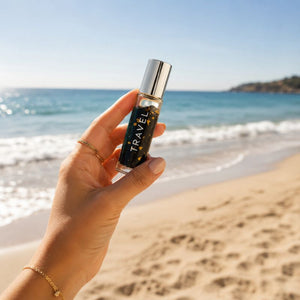 Hand holding a small bottle labeled 'TRAVEL' on a beach with ocean and sky in the background