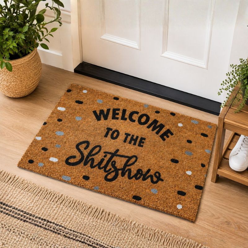 A brown coir doormat with the text 'WELCOME TO THE Shitshow' in black, placed in front of a white door on a wood floor with potted plants and a small wooden side table nearby.-Welcome to the Shitshow Doormat Black